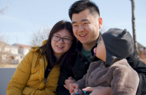 Kaiwen Xu smiling, outside during the winter, with his arms around his wife and son.