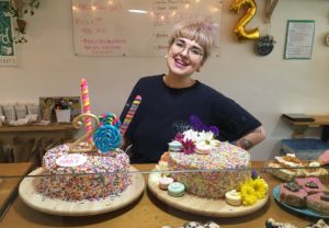 Jo Masterson stands smiling in front of two delightfully sprinkled cakes