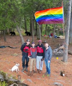 A group stands with Adam beside a pride flag at a campground near the water. 