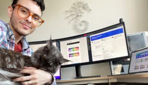 Jesse Pyne, Founder of Purpose Fuel sits in front of his computer, holding his grey cat and smiling. 