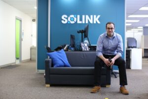 The founder of Solink sits on the edge of a small couch, leaning forward with hands folded, in front of a blue feature wall with the Solink logo on it. 