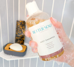 A person extends their soapy hand out of the shower, holding a shampoo bottle that reads The Better Scalp Company