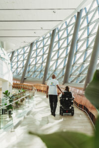 A person walks along the curved window of the PROOF gallery, alongside someone in a wheelchair. 