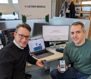 The founders of Leynek Medical sit with computer monitors behind them. One is holding a phone showing the company's app. 
