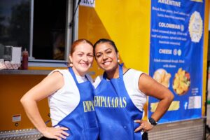 Two women stand together smiling in front of a food operation wearing blue aprons with Empanada's written in yellow. 