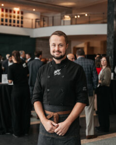 Owner of My Catering Group Derick Cotnam stands wearing a black, branded chefs coat in an event space. 