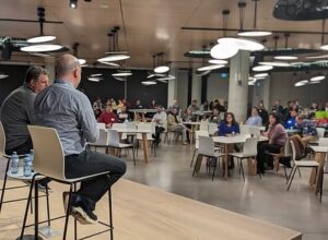 Image: Two individuals seated and on stage address the crowd at an event