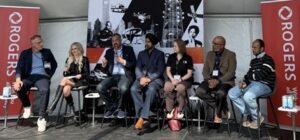 A panel is gathered on stage, seated on high table chairs at an event. 