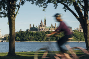 A cyclist zooms by the camera while riding along the Ottawa River with Parliament in the background. 
