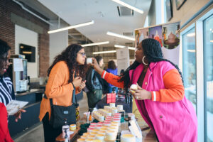 A vendor interacts with a customer at an indoor marketplace, sharing the scent of one of her products.