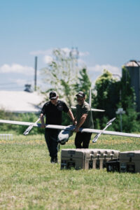 Two people carry out a large drone across a grassy field on a sunny day.