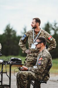 Military personnel in uniform work in tandem to perform drone exercises. One is standing, the other is seated at a small desk, with virutal glasses operating a drone. 
