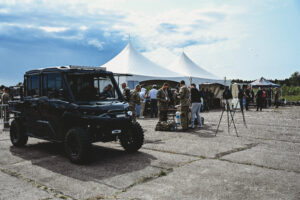 A tactical vehicle is parked on an old runway that is hosting an event, with two visible tents and people congregating around the area.
